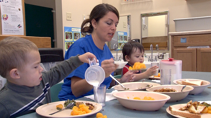 A staff member sits with two toddlers teaching buffet style family dining