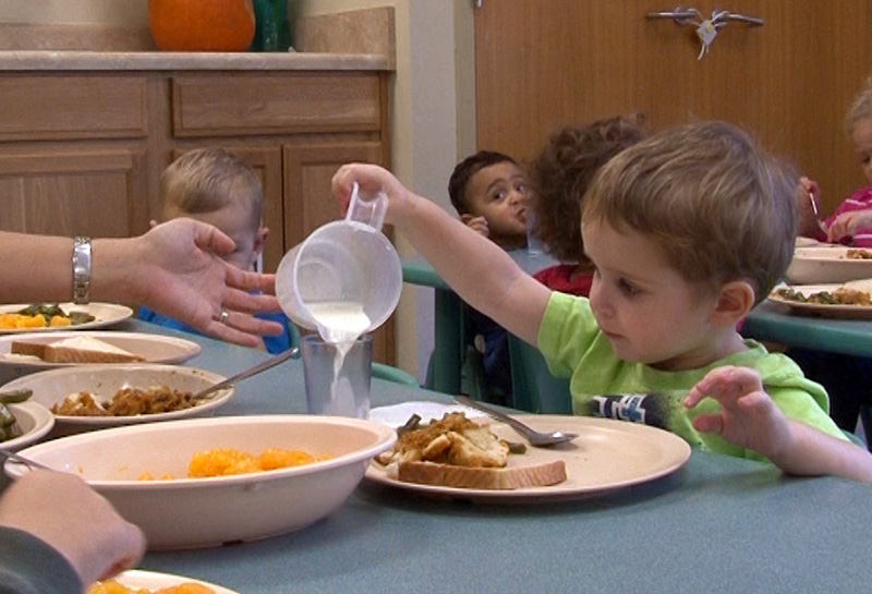 A staff member assists a toddler as he tries pouring milk into his glass during family-style dining
