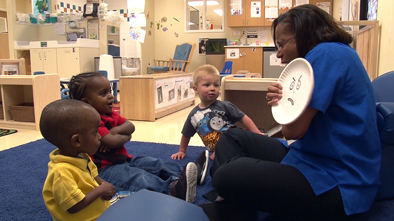 A caregiver holds up facial expressions on a paper plate while explaining and demonstrating different emotions