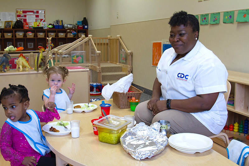 A caregiver sits with two toddlers at a table during meal-time.