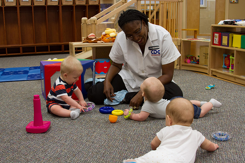 A caregiver sits and plays with three infant-toddlers on the floor