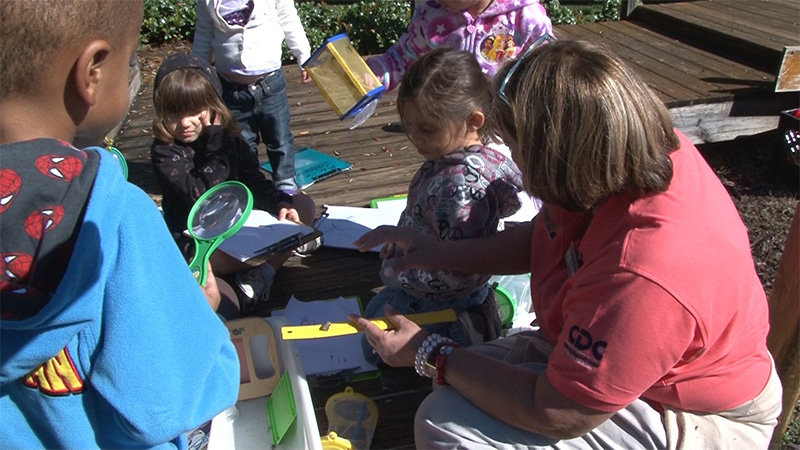 A care-giver sits with children outside as they explore bugs with magnifying glasses