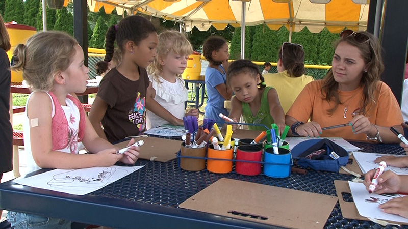 Children and a caregiver color with markers while sitting around a table outside